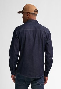 Camisa de mezclilla azul oscura de manga larga, con cuello estándar y puños abotonados. Combinada con jeans azul oscura y una gorra de béisbol color beige.