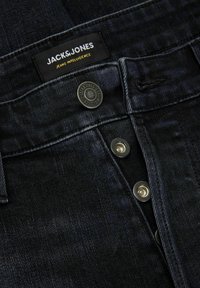 Jeans de mezclilla oscura con una vista en primer plano de la cinturilla, que presenta una etiqueta de marca negra, un botón de metal y un detalle de costura visible.