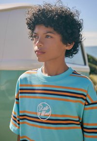 Pruhované tričko v tyrkysové barvě, s oranžovými a tmavě modrými pruhy. Bílé kruhové logo s textem "Skate or Mind" na hrudi. Krátké rukávy.