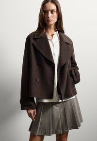 Bruine wolmix dubbelzijdige jas met grote revers en paarse knopen. Gecombineerd met een witte blouse en een grijze geplooide rok.