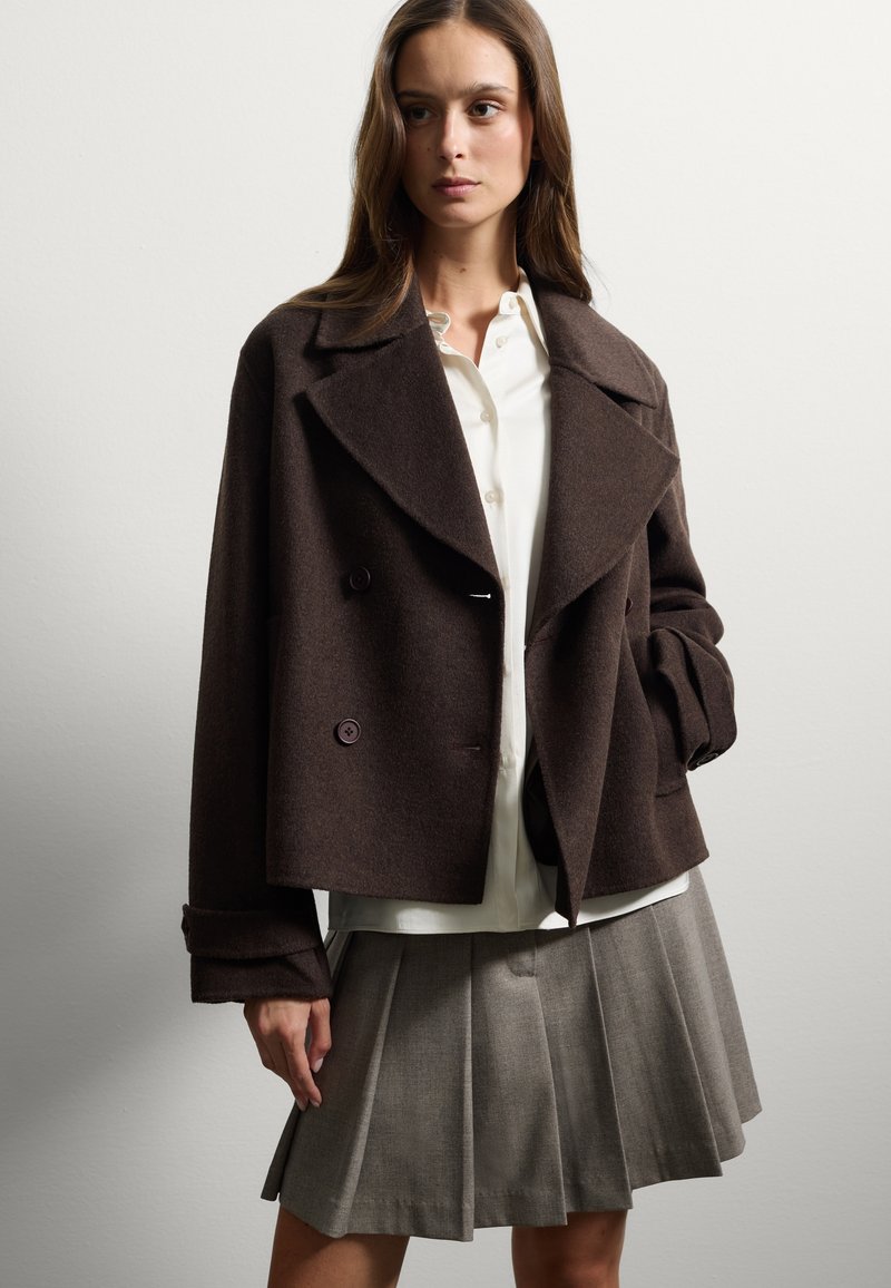 Bruine wolmix dubbelzijdige jas met grote revers en paarse knopen. Gecombineerd met een witte blouse en een grijze geplooide rok.