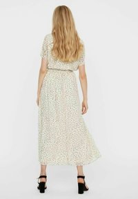 Robe longue crème avec des taches noires et vertes, manches courtes, ceinture élastique et tissu fluide. Associée à des sandales à talons noires.