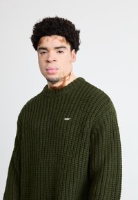 Σκούρο πράσινο πλεκτό πουλόβερ με υφή και στρογγυλή λαιμόκοψη. Μικρό λευκό λογότυπο "OBEY" μπροστά. Χαλαρή εφαρμογή.