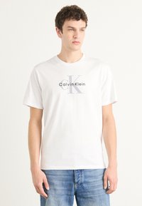 Bijela pamučna majica s okruglim izrezom, siva "Calvin Klein" oznaka i stilizirani "CK" grafički prikaz na prednjoj strani.