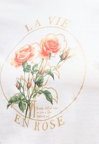 Δύο ροζ τριαντάφυλλα με πράσινα φύλλα μέσα σε έναν χρυσό κύκλο που περιβάλλει το κείμενο «LA VIE EN ROSE» σε λευκό φόντο.