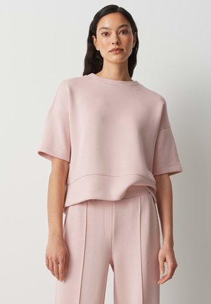 Woman with long dark hair wearing loose light pink short-sleeve top and matching pants, standing against a plain light background.