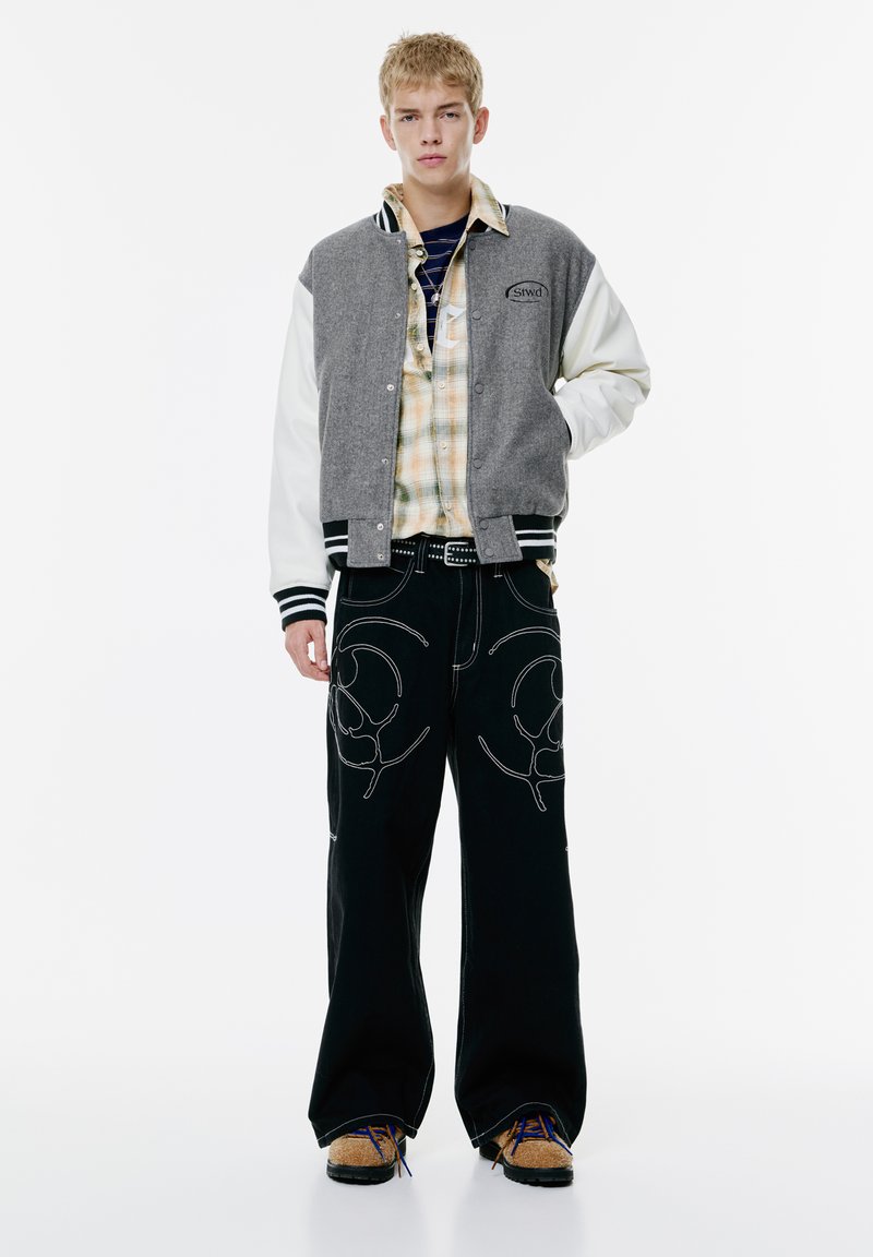Veste de varsity gris et blanche avec des poignets rayés, chemise à carreaux en dessous, large pantalon noir avec broderie blanche, et chaussures marron.