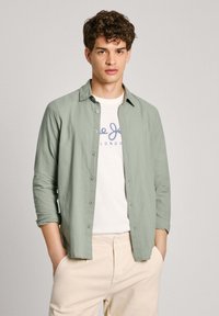 Camicia button-up verde chiaro realizzata in un tessuto morbido, con colletto, maniche lunghe e bottoni frontali, indossata sopra una t-shirt bianca con logo.