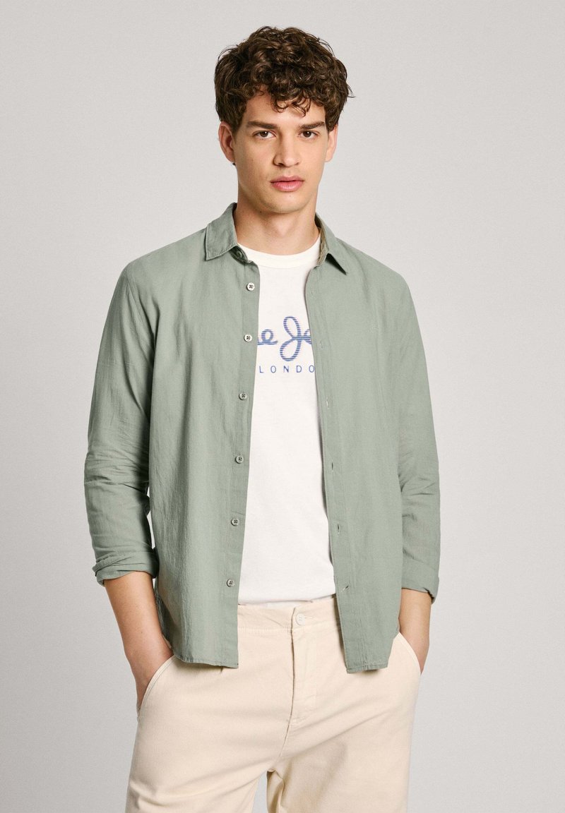 Camicia button-up verde chiaro realizzata in un tessuto morbido, con colletto, maniche lunghe e bottoni frontali, indossata sopra una t-shirt bianca con logo.