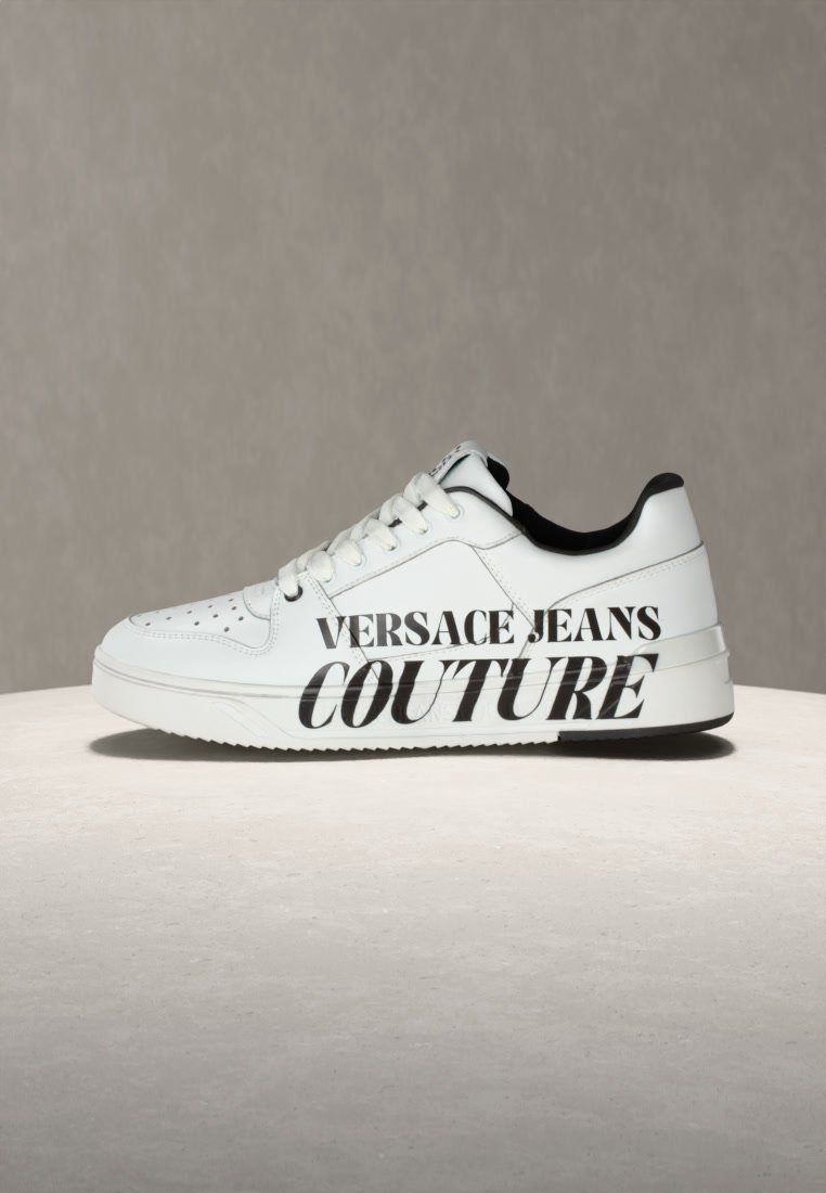 Бял нисък кец с черен вътрешен подгъв и голям черен текст „"Versace Jeans Couture"" отстрани, показан на неутрален фон.