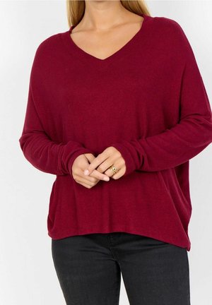 Femme portant un pull bordeaux à col en V manches longues et un pantalon noir, les mains légèrement croisées à la taille, montrant une bague en or.