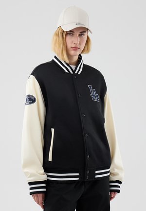 Ung person med blondt hår bærer en sort og hvid LA Dodgers varsity jakke og en lyse beige baseballkasket, stående mod en ensfarvet baggrund.