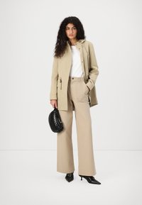 Giacca in cotone beige con chiusura a toggle, abbinata a pantaloni beige a gamba larga. Una borsa nera textured e décolleté nere a punta completano il look.