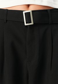 Svarta byxor med en slät tygkvalitet, en fyrkantig silverfärgad bältesspänne och frampocket. En enkel och skräddarsydd design.
