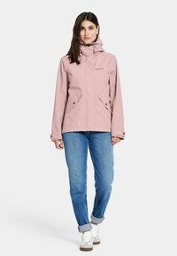 Lichtroze waterdichte jas met een capuchon, ritssluiting, twee voorzakken en verstelbare manchetten, gecombineerd met blauwe jeans en witte sneakers.