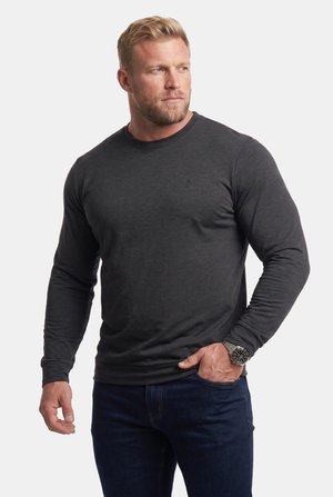 Blonder Mann mit Bart, der ein dunkelgraues Langarmshirt, dunkle Jeans und eine große Armbanduhr trägt und mit einer Hand in der Tasche zur Seite schaut.