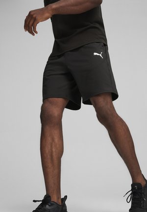 Pantalones cortos atléticos negros de tela suave, con un ajuste holgado y un logo blanco de Puma en el lado izquierdo. Combinados con zapatillas negras.