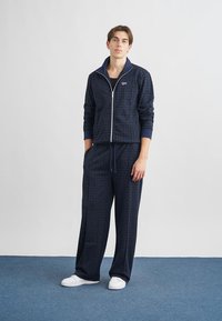 Tumši zils sporta tērps no mīksta materiāla ar rakstainu dizainu un logotipu, rāvējslēdzēja jaka un brīva piegriezuma bikses. Valkātas baltas sporta kurpes.