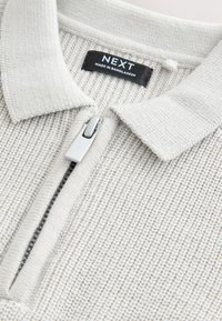 Hellgrauer Pullover mit Reißverschluss und strukturiertem Strickmuster, mit Polokragen und silbernem Reißverschluss. Sichtbares Markenetikett: NEXT, hergestellt in Bangladesch.