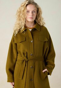 Manteau en mélange de laine vert olive avec une taille ceinturée, de grandes poches avant, une fermeture à boutons et un col. Texture douce avec une coupe décontractée.