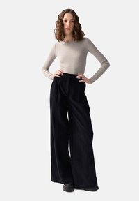 Jasnoszara bluzka z długim rękawem w połączeniu z czarnymi, szerokimi spodniami z korduroyu o wysokim stanie. Modelka stoi swobodnie, z rękami na biodrach, na jasnym tle.