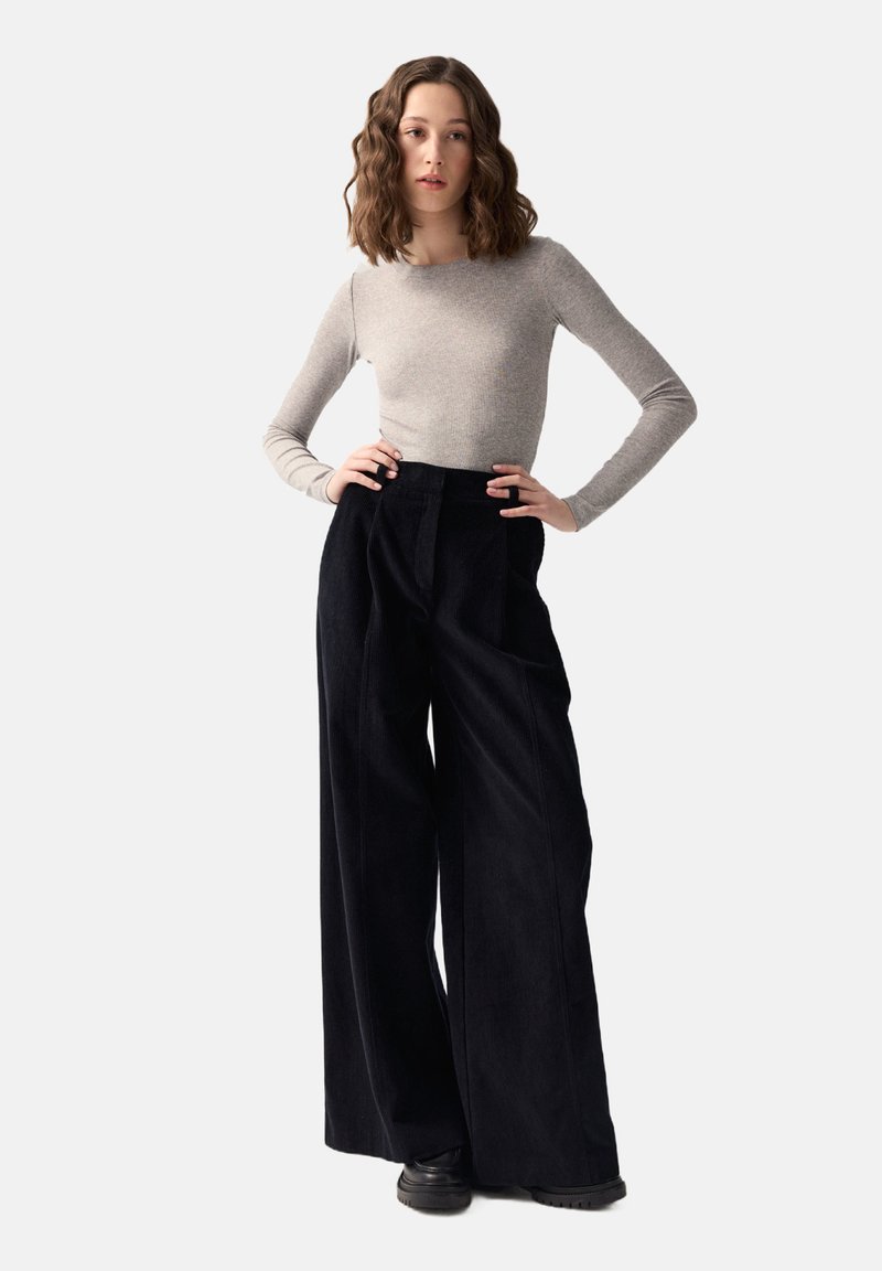 Jasnoszara bluzka z długim rękawem w połączeniu z czarnymi, szerokimi spodniami z korduroyu o wysokim stanie. Modelka stoi swobodnie, z rękami na biodrach, na jasnym tle.