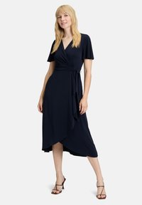 Robe portefeuille en tissu bleu marine avec des manches courtes à volants, un décolleté en V et un hem asymétrique. Texture lisse, fermeture à nouer réglable à la taille.