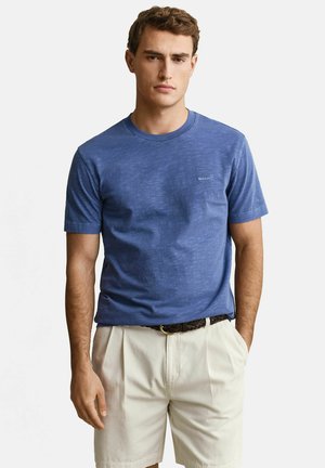 Jonge man met een blauw T-shirt met korte mouwen en lichte beige korte broek met een donkere gevlochten riem, staand met één hand in zijn zak.