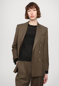 Olijfgroene op maat gemaakte blazer met dubbele knopen, inkepingen op de revers en subtiele schoudervulling, gedragen over een zwarte textuur top.