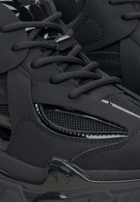 Schwarze Sportschuhe aus einer Kombination von glattem synthetischem Leder und Netzgewebe, mit flachen schwarzen Schnürsenkeln, strukturierten Akzenten und einem eleganten Design.