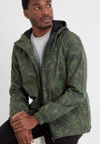Green camouflaged lightweight jacket in a shiny fabric, featuring a zip closure, adjustable hood, and elastic cuffs. The design includes abstract patterns.