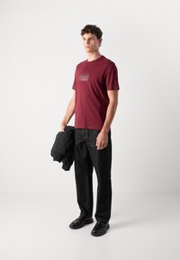 Bordo spalvos medvilnės marškinėliai su "HUGO" logotipu, trumpomis rankovėmis, derinami su tamsiomis džinsinėmis kelnėmis ir juodais batais. Modelis laiko juodą švarką.