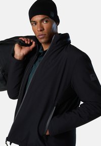 Giacca soft shell nera con collo alto, tasche con cerniera e tessuto strutturato. Indossata con un berretto nero e una borsa.