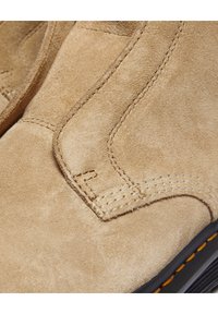 Gros plan d'une botte en daim beige montrant les coutures détaillées et une partie de la semelle noire avec des accents de couture orange.