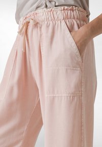 Lichtroze, losse broek met een elastische tailleband, decoratieve strikjes en twee zijzakken. Gemaakt van zachte, gestructureerde stof.