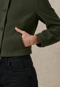 Green, wool-blend cropped jacket with a collar, black buttons, and a side pocket; worn over dark denim jeans.