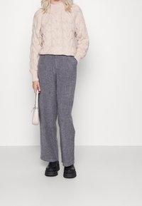 Femme portant un pull en maille torsadée rose clair, un pantalon large gris, des bottes à plateforme noires, et tenant un petit sac à main beige.