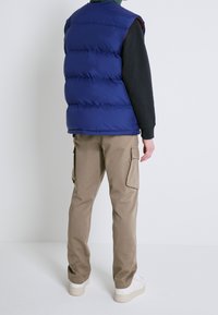 Blaue ärmellose Steppweste mit gesteppte Abschnitte, getragen über einem schwarzen Langarmshirt, kombiniert mit beigen Cargohosen und weißen Turnschuhen.