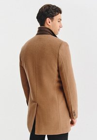 Brązowy wełniany płaszcz z wysokim kołnierzem, dopasowany krój oraz akcenty trzech guzików na mankietach. Gładka faktura, płaszcz o długości do połowy uda oraz wentylowane plecy.