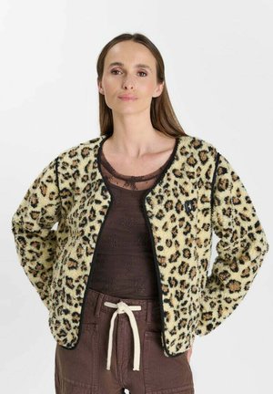 Frau mit langen braunen Haaren trägt eine Fleecejacke mit Leopardenmuster über einem transparenten dunklen Top und braunen Hosen mit weißem Kordelzug.
