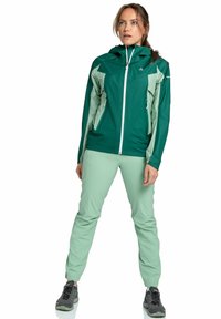 Zielona i jasnazia zielona sportowa kurtka z kapturem, zamek błyskawiczny z przodu i dwoma bocznymi kieszeniami. Połączona z jasnazi zielonymi spodniami i ciemnymi butami.