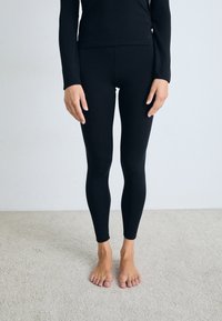 Persona in piedi a piedi nudi su un tappeto, indossando leggings neri e una maglietta nera a maniche lunghe, davanti a una parete grigia chiara e semplice.