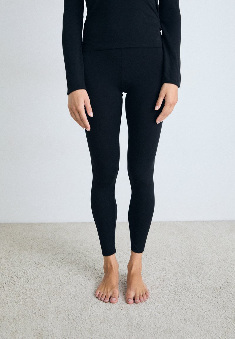 Persona in piedi a piedi nudi su un tappeto, indossando leggings neri e una maglietta nera a maniche lunghe, davanti a una parete grigia chiara e semplice.