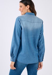 Camicia in denim azzurro chiaro con maniche lunghe, polsini a bottone e dettaglio sulla spalla posteriore, caratterizzata da una texture leggermente sbiadita per un aspetto morbido.