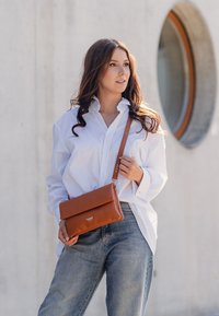 Hnědá kožená crossbody taška s klopou a embosovaným logem, nošená s bílou oversized košili a světle modrými džíny.