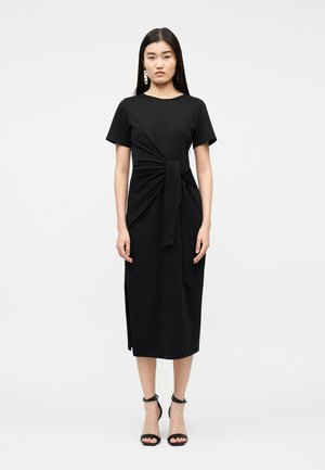 Femme aux longs cheveux noirs portant une robe midi noire à manches courtes, avec un nœud sur le côté, et des talons noirs à bride à la cheville, debout sur un fond uni.