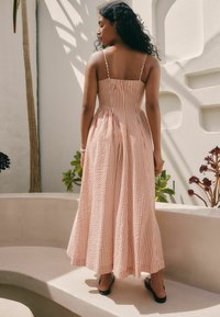 Robe maxi en rose clair avec des rayures blanches alternées. Elle possède un corsage ajusté, des bretelles spaghetti réglables et une jupe fluide.