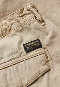 Beige stof met een subtiele textuur, voorzien van een zwart label met de tekst "SUPERDRY" in gouden letters en een gestikt pocketdetail.
