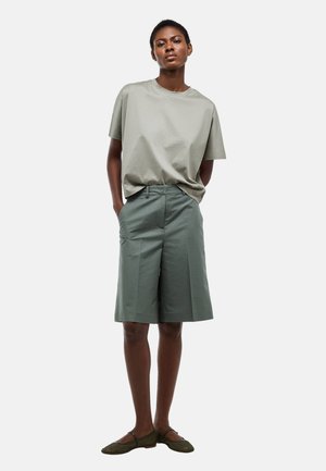 Femme debout, mains dans les poches, portant une chemise ample à manches courtes gris clair, un short gris-vert au genou et des ballerines vertes, sur fond blanc.