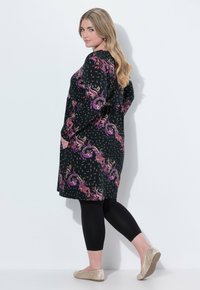 Robe noire avec un motif paisley violet vif, manches longues, poches latérales, portée avec des leggings noirs et des chaussures dorées à enfiler.